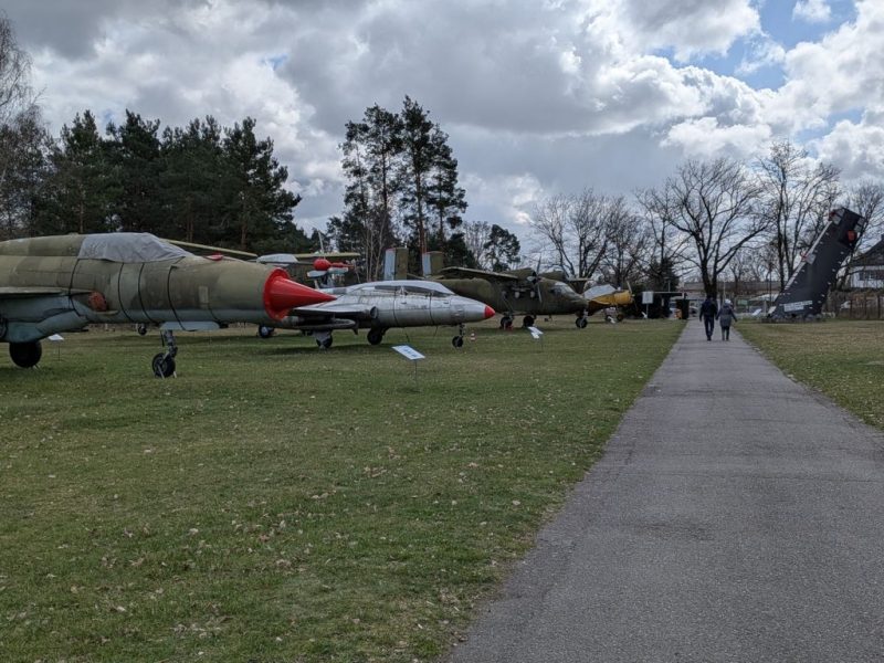 Historische Militärflugzeuge in Grau und Metalltönen stehen auf einer offenen Wiese, Baumreihe im Hintergrund; ein Weg führt Besucher vorbei