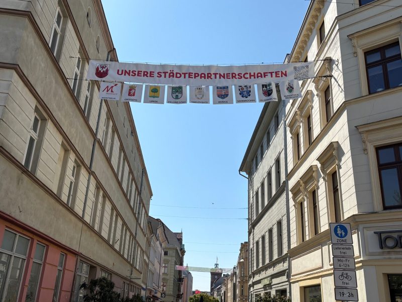 Über der Sprem hängt ein Banner mit unseren Städtepartnerschaften und den Wappen von Montreuil, Grosseto, Zielona Góra und weiteren Städten.