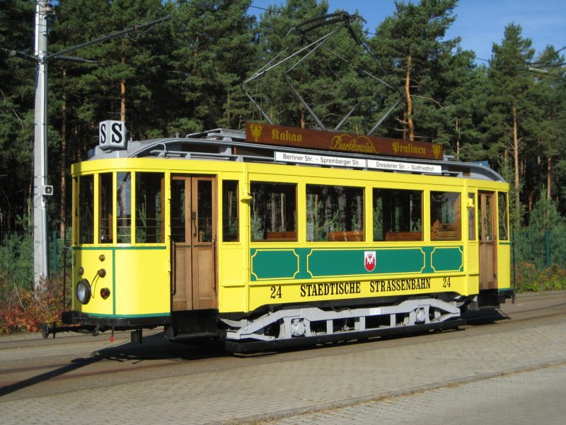 Eine historische gelb-grüne Straßenbahn der Städtischen Straßenbahn Cottbus steht an einem Waldstück, mit Zieltafeln und Pantograph unter freiem Himmel.