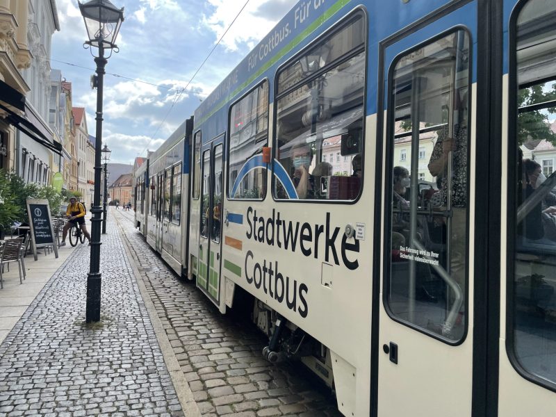 Eine blaue und weiße Straßenbahn der Stadtwerke Cottbus fährt durch eine historische Kopfsteinpflasterstraße, während Fahrgäste durch die Fenster blicken.