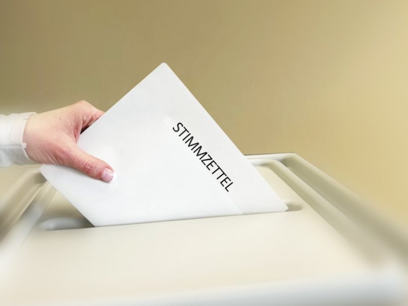 Eine Hand wirft einen Stimmzettel in eine Wahlurne, während das Wahlpapier halb in den Schlitz gleitet und der Wahlvorgang beginnt.