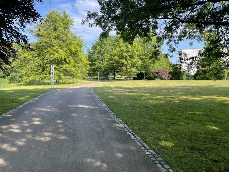 Ein geschwungener Weg führt durch eine grüne Parkanlage mit großen Bäumen, gepflegten Rasenflächen und einem hellen Gebäude im Hintergrund.