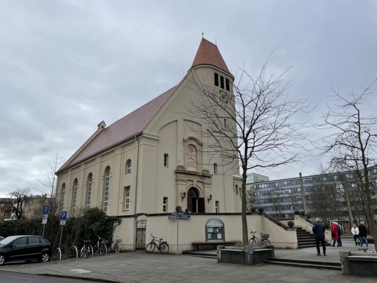 Die Lutherkirche in Cottbus zeigt einen hellen Kirchenbau mit rotem Dach, Turm mit Uhr und spitzem Helm an einem grauen Tag.