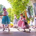 Beim sorbischen Fest in Cottbus tanzen Kinder in traditionellen Trachten auf einer Bühne vor Publikum unter sommerlichen Bäumen beim Stadtfest.