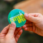 Hände halten einen türkisfarbenen Button mit der Aufschrift „COTTBUS OPEN“ und „interkulturelles Festival“ beim Cottbuser Stadtfest.