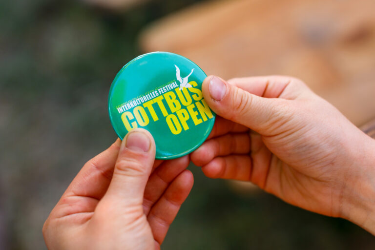 Hände halten einen türkisfarbenen Button mit der Aufschrift „COTTBUS OPEN“ und „interkulturelles Festival“ beim Cottbuser Stadtfest.