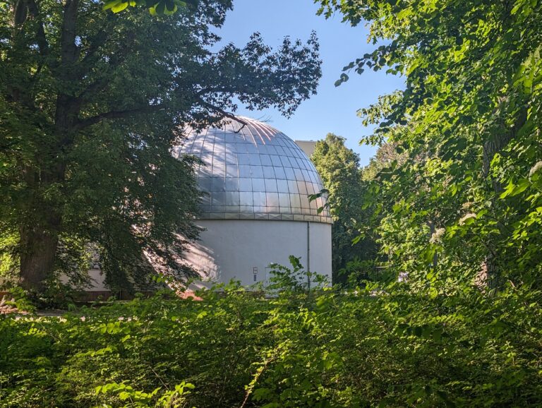 Eine silberne Kuppel eines Gebäudes ragt zwischen dichtem Grün und hohen Bäumen unter blauem Himmel hervor.