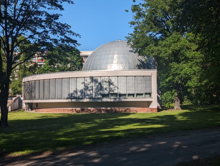 Rundes Planetarium mit silberner Kuppel in einer grünen Parkanlage, umrahmt von hohen Bäumen bei Sonnenschein.