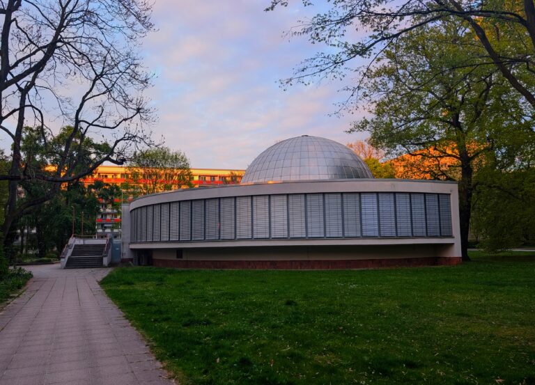 Rundes Gebäude mit gläserner Kuppel im Park, umgeben von Bäumen und einem Weg im Abendlicht dort.
