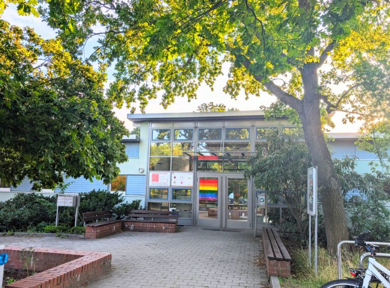 Eingang eines Sozialkulturellen Zentrums mit Regenbogenflagge, Glasfassade und großem Baum im Vordergrund.