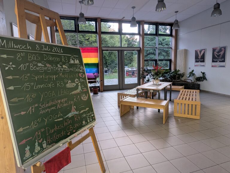 Ein heller Raum mit Kreidetafel, Tagesplan, Regenbogenflagge am Fenster, Holzbänken und Pflanzen.