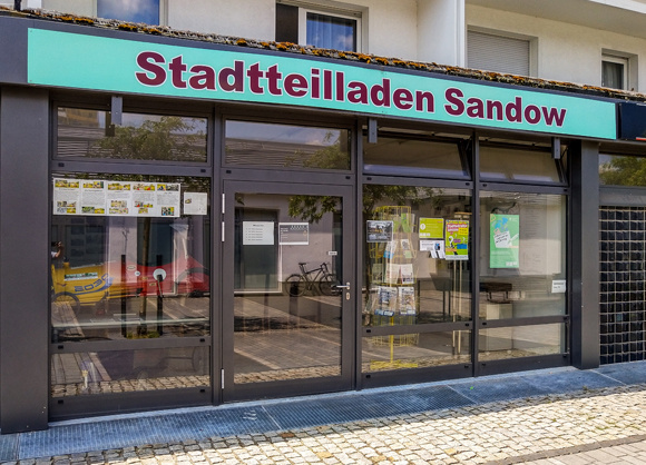 Der Stadtteilladen Sandow mit großer Schaufensterfront und Eingangstür an einer gepflasterten Straße.
