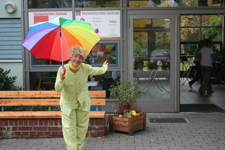 Eine Frau in hellgrüner Kleidung steht vor dem Soziokulturellen Zentrum und hält einen Regenbogen-Regenschirm.