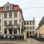 Historisches Gebäude mit rotem Ziegeldach, großen Fenstern und einem Eingangstor an einer schmalen Straße.