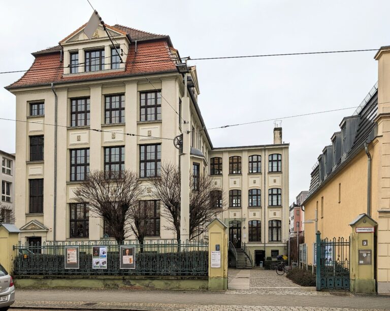 Historisches Gebäude mit rotem Ziegeldach, großen Fenstern und einem Eingangstor an einer schmalen Straße.