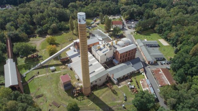 Luftaufnahme einer Industrieanlage mit hohem Schornstein, mehreren Gebäuden und Zufahrten am Rand eines Waldgebiets.