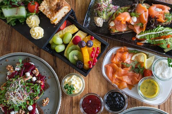 Auf dem Holztisch liegen Räucherlachs, Obstsalat, Blattsalat und mehrere Dips neben kleinen Speisen und frischen Kräutern.