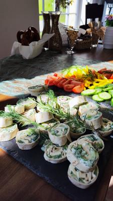 Auf einem Buffet liegen gerollte Häppchen, frisches Gemüse, Kräuter und belegte Röllchen neben Brot und Obst.