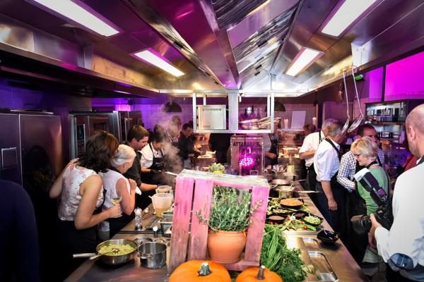 Mehrere Gäste und Köche stehen in einer beleuchteten Küche an Buffets und Kochstationen zwischen dampfenden Speisen.