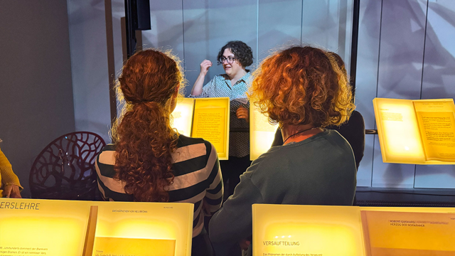 Drei Besucherinnen stehen vor beleuchteten Ausstellungstafeln, während eine Frau hinter Glas erklärt.