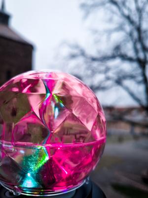 Eine pinke Spiegelkugel mit bunten Lichtreflexen steht im Freien; dahinter sind unscharf Bäume und ein Gebäude zu sehen.