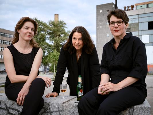 Drei Frauen in schwarzer Kleidung sitzen auf einer Steinbank; vor ihnen stehen ein Glas und eine Bierflasche.