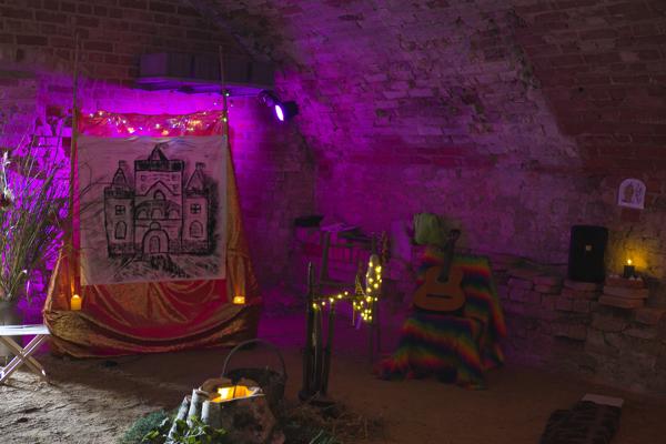 Violettes Licht fällt in einen Gewölberaum mit Draperie, Hauszeichnung, Kerzen, Lichterkette und bunt verhülltem Instrument.