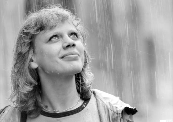 Eine junge Frau blickt im Regen nach oben, während Tropfen über ihr Gesicht und ihre Haare laufen.