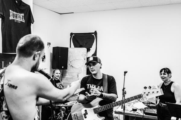 Mehrere Musiker proben in einem kleinen Raum, während ein Bassist im Vordergrund spielt und eine Sängerin am Mikrofon steht.