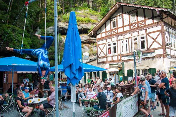 Eine blau gekleidete Akrobatin hängt kopfüber an Tüchern vor einem Fachwerkhaus mit Gästen und Sonnenschirmen.