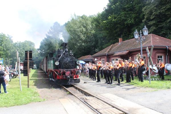 Eine schwarze Dampflok fährt an einer Station vorbei, während eine Blaskapelle in Orange und Schwarz daneben aufgestellt ist.