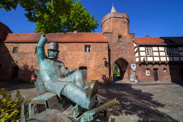 Vor einem Backsteintor mit Turm sitzt eine Bronzefigur mit Zylinder, erhobener Hand und ausgestrecktem Bein.