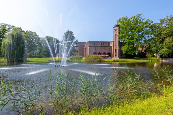 Ein Springbrunnen sprudelt in einem Teich vor einem historischen Backsteingebäude, umgeben von Bäumen und Ufergras.