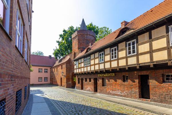 Eine schmale Altstadtgasse mit Kopfsteinpflaster führt an Fachwerk- und Backsteinbauten vorbei, neben einem runden Turm.