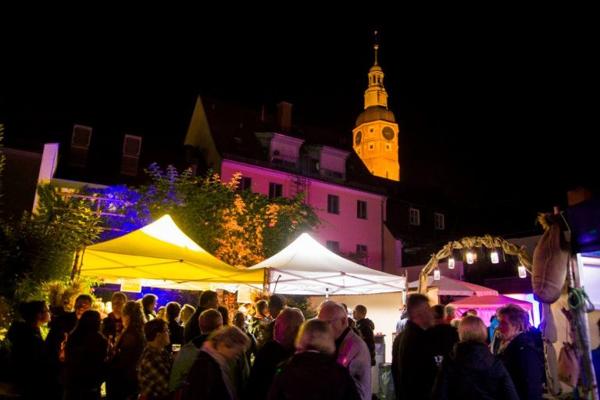 Bei Nacht drängen sich viele Menschen zwischen gelben und weißen Zelten; im Hintergrund leuchtet der Kirchturm.