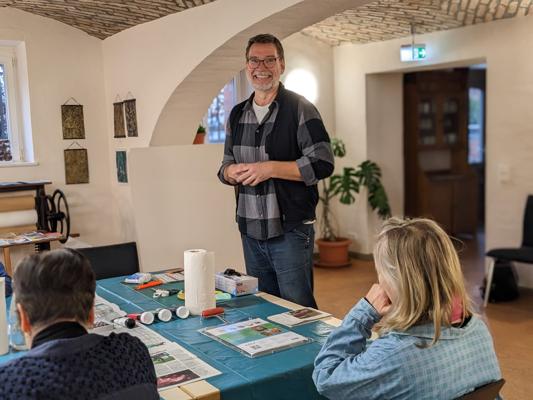 Ein lächelnder Mann steht in einem Raum mit Gewölbedecke vor einem Tisch mit Druckmaterialien und Papieren.