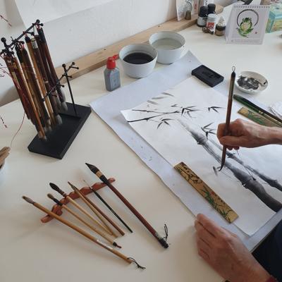 Eine Hand malt mit einem Pinsel ein Bambusmotiv in Tusche auf Papier; daneben liegen mehrere Pinsel, Tusche und Schalen.