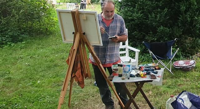 Ein Mann arbeitet im Grünen an einer Staffelei und hält eine Palette; daneben stehen Tisch und Malutensilien.