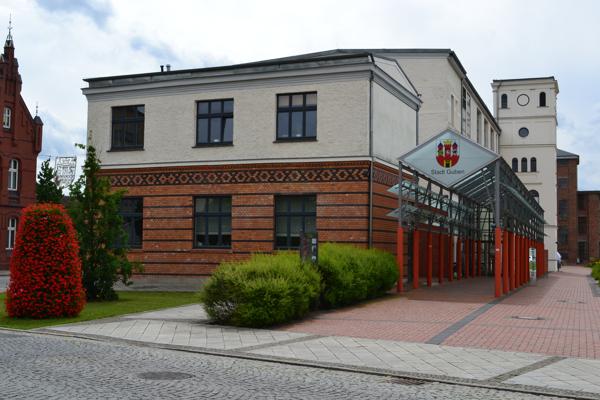 Am Gebäude mit dem Schild „Stadt Cottbus“ führt ein Glasdach zum Eingang; davor liegen Pflasterweg, Grünstreifen und Büsche.