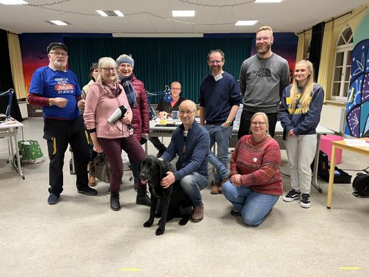 Mehrere Helfende stehen und sitzen in einem Saal, während ein schwarzer Hund vorn zwischen ihnen sitzt.