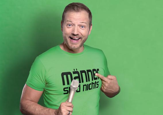Ein lachender Mann im grünen T-Shirt hält ein Mikrofon und zeigt auf die Aufschrift „Männer sind nichts“.