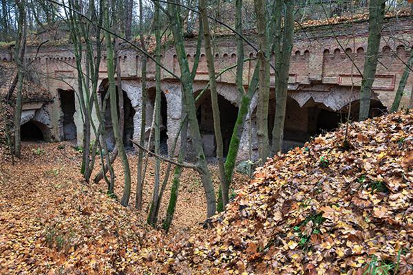 Eine alte Ziegelbefestigung mit Bögen und Öffnungen steht zwischen kahlen Bäumen, davor liegen Hügel aus herbstlichem Laub.