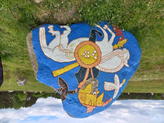 Ein bemalter Findlingsstein zeigt auf blauem Grund stilisierte Tiere, Figuren und ein rundes Zentrum mit farbigen Mustern.