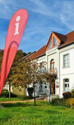 Vor einem weißen Museumsgebäude steht eine rote Infofahne, daneben wachsen Bäume und ein gepflegter grüner Garten.