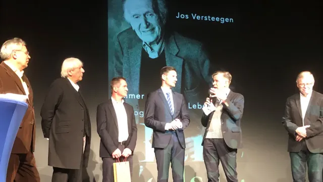 Sechs Männer stehen auf einer beleuchteten Bühne vor einer großen Leinwand, auf der ein Porträt und Text eingeblendet sind.