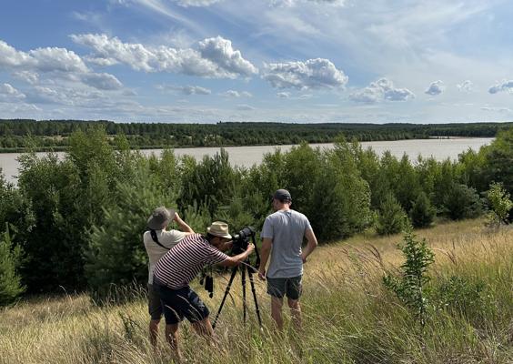 Drei Personen stehen auf einer grasigen Anhöhe mit Kamera und Stativ und blicken über ein Seeufer mit Wald und Wolkenhimmel.