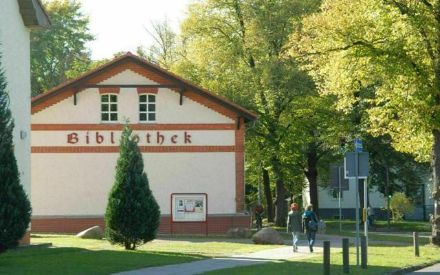 Vor einem Bibliotheksgebäude mit dem Schriftzug „Bibliothek“ gehen zwei Personen über einen Weg zwischen Bäumen.