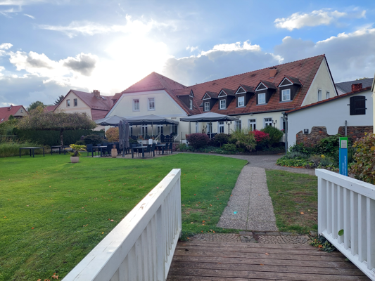 Vor einem hellen Haus mit rotem Dach liegen eine gepflegte Rasenfläche, eine Terrasse und ein kleiner Steg.