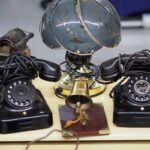 Zwei schwarze Wählscheibentelefone stehen neben einer dekorativen Tischlampe mit blauem Schirm und kleiner Glocke.