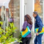 Vor einem Schaufenster mit Mantel stehen zwei Personen und schauen hinein; beide tragen blaue Cottbus-Einkaufstaschen.